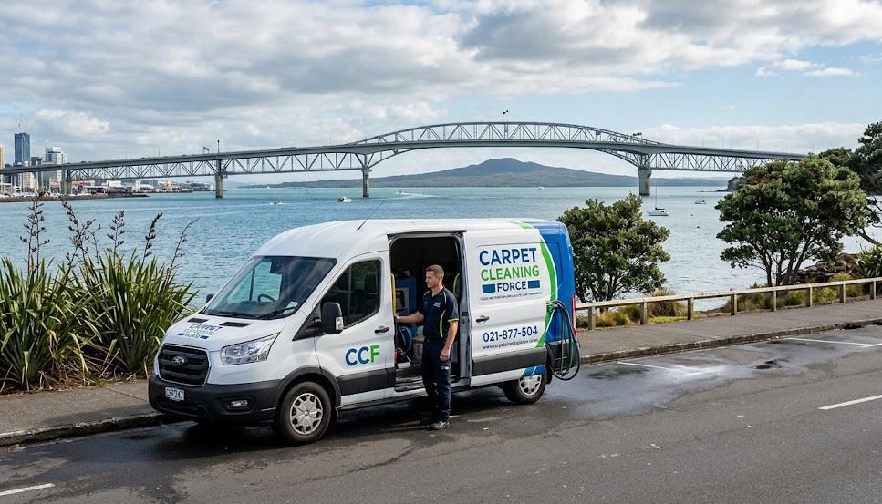 A photographic hero image showing a white Carpet Cleaning Force (CCF) truck-mount van, branded as "Flood Restoration Specialists," parked on a scenic coastal road in Devonport, North Shore, Auckland. In the background, the Auckland Harbour Bridge spans the harbour, with Rangitoto Island centered prominently behind it, providing local North Shore authority proof.